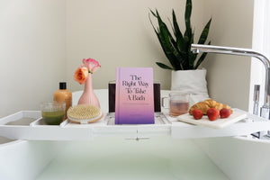 The Right Way To Take A Bath book on white bath caddy over luxury white bathtub. White bath caddy holding green juice in vintage style drinking glass, fall themed bubble bath, dry brush for exfoliation, tea in iridescent mug, white marble cheeseboard holding strawberries and croissant with a snake plant in the bathroom.