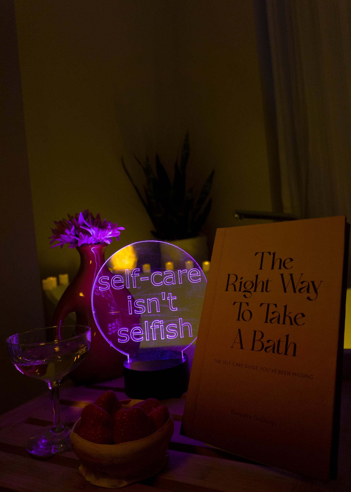 Light up sign for room with a self care message in front of bathtub with candles next to glass of champagne and strawberries and The Right Way To Take A Bath book.