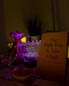 Light up sign for room with a self care message in front of bathtub with candles next to glass of champagne and strawberries and The Right Way To Take A Bath book.