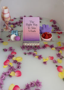 Milk bath with flowers with wire bath caddy holding pink marble dish, blue bubble candle, purple bath bomb, sisal towel for exfoliation, The Right Way To Take A Bath book, purple exfoliating gloves and strawberries for bubble bath.