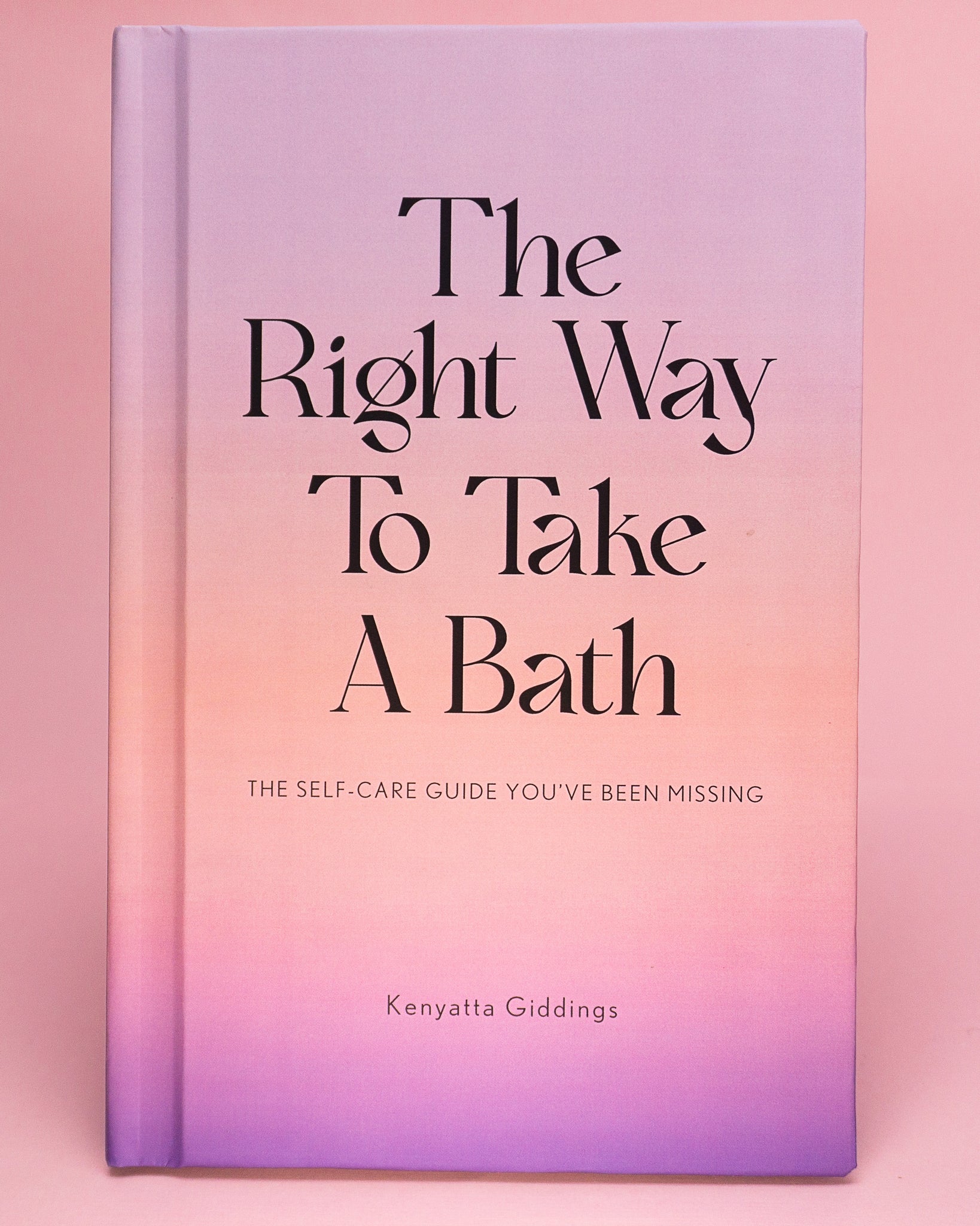 The Right Way To Take A Bath hardcover book with a soft touch matte pastel cover against a pink background. The Right Way To Take A Bath is a self-care guide that captures bubble bath tips in a decor-friendly book that makes the perfect spa gift.